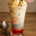 A tall glass of Royal Falooda dessert layered with rose syrup, basil seeds, sev, sweetened milk, topped with vanilla ice cream, nuts, and tutti frutti.